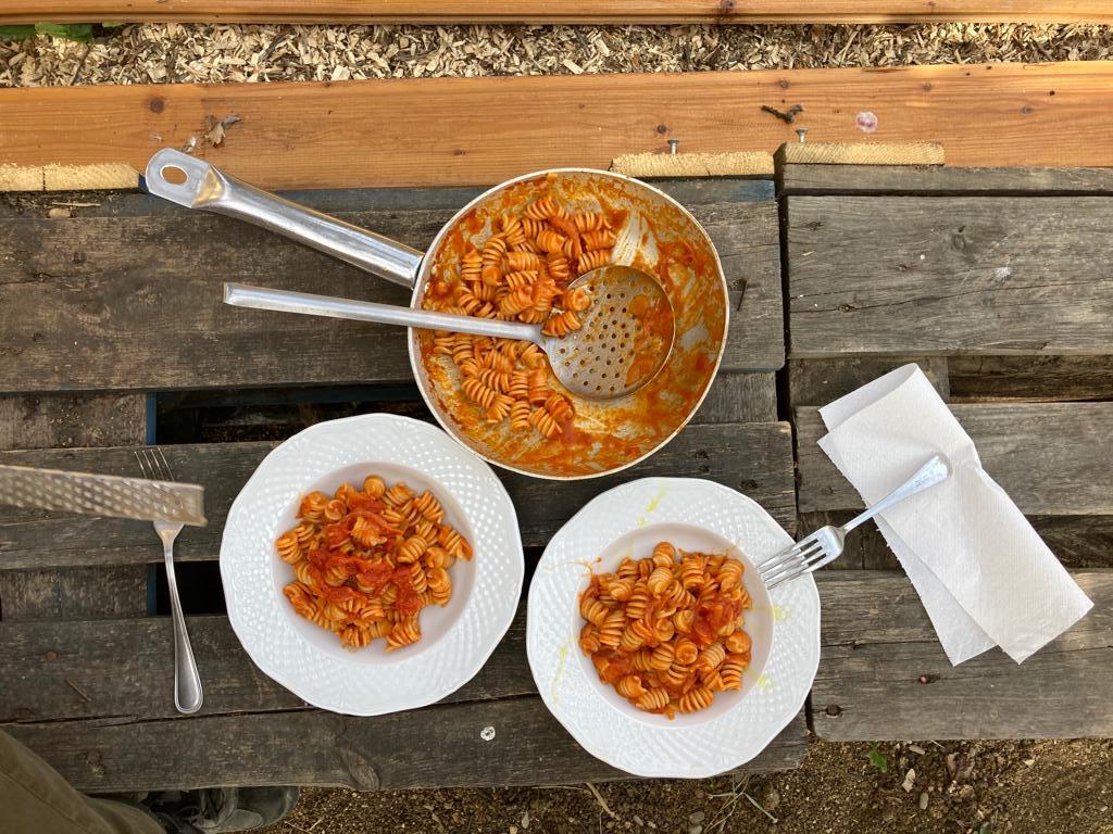 A pan of curly pasta with tomato sauce and a serving spoon next two two bowls of the pasta. They are set on a rustic wood table, outside.