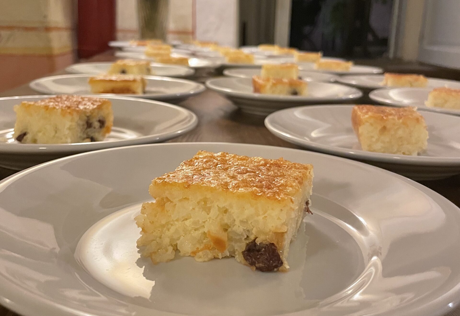 A tableful of plates, each with one square of "dolce di riso." In the foreground is a close up piece of the cake, with a gently browned top, grains of rice and speckled with raisins.