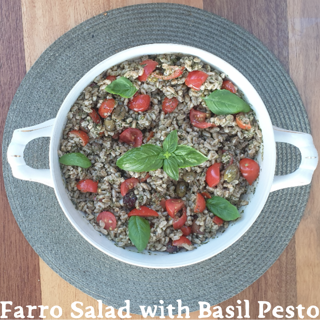 A white ceramic serving bowl filled with farro, chopped tomatoes, basil, and green olives. Garnished with fresh basil and set on a woven placemat and wood table. "Farro Salad with Basil Pesto" is written at the bottom.