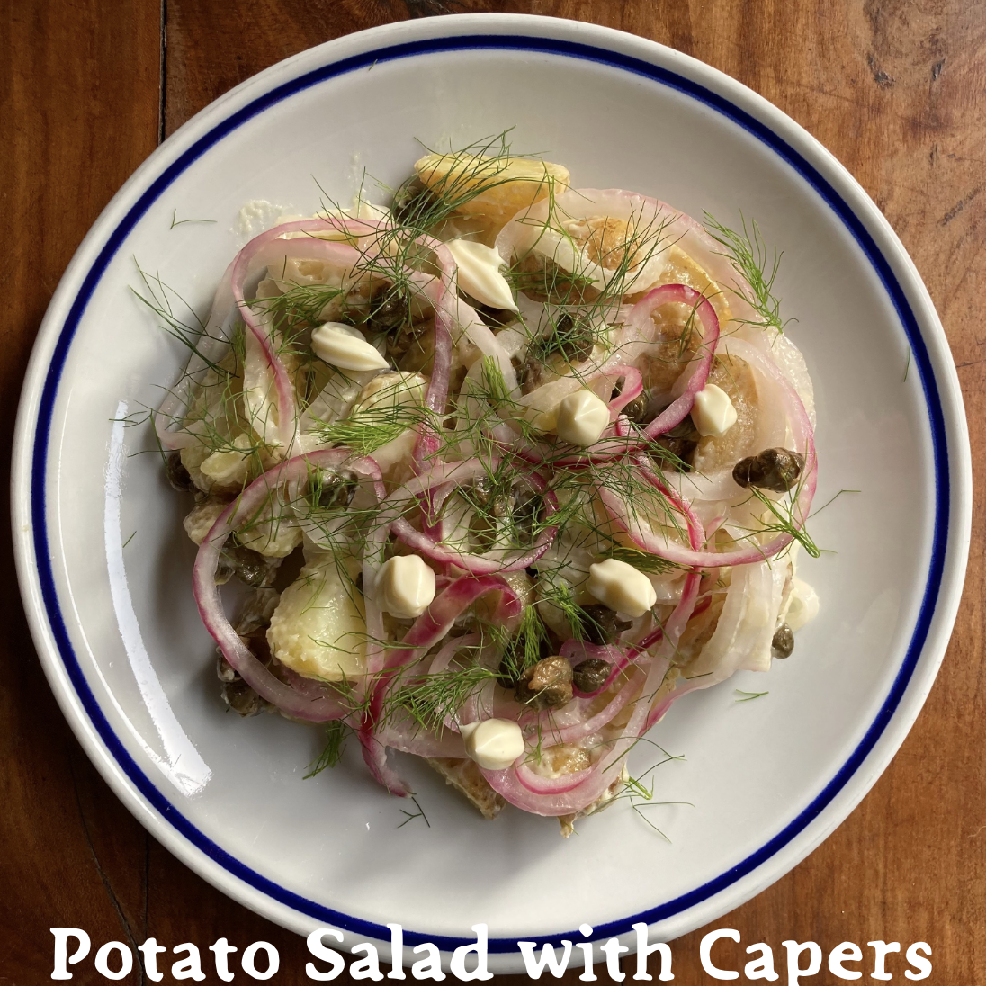 A white ceramic plate on wooden table surface, layered with wedges of potatoes, pickled red onions, capers, dollops of mayonnaise, and fresh fennel fronds. "Potato Salad with Capers" is written at the bottom.