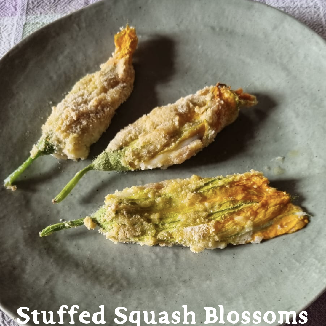 Three lightly breaded and baked vibrant zucchini flowers on a rustic gray ceramic plate, on a light green and lavender checked tablecloth. "Stuffed Squash Blossoms" is written at the bottom.