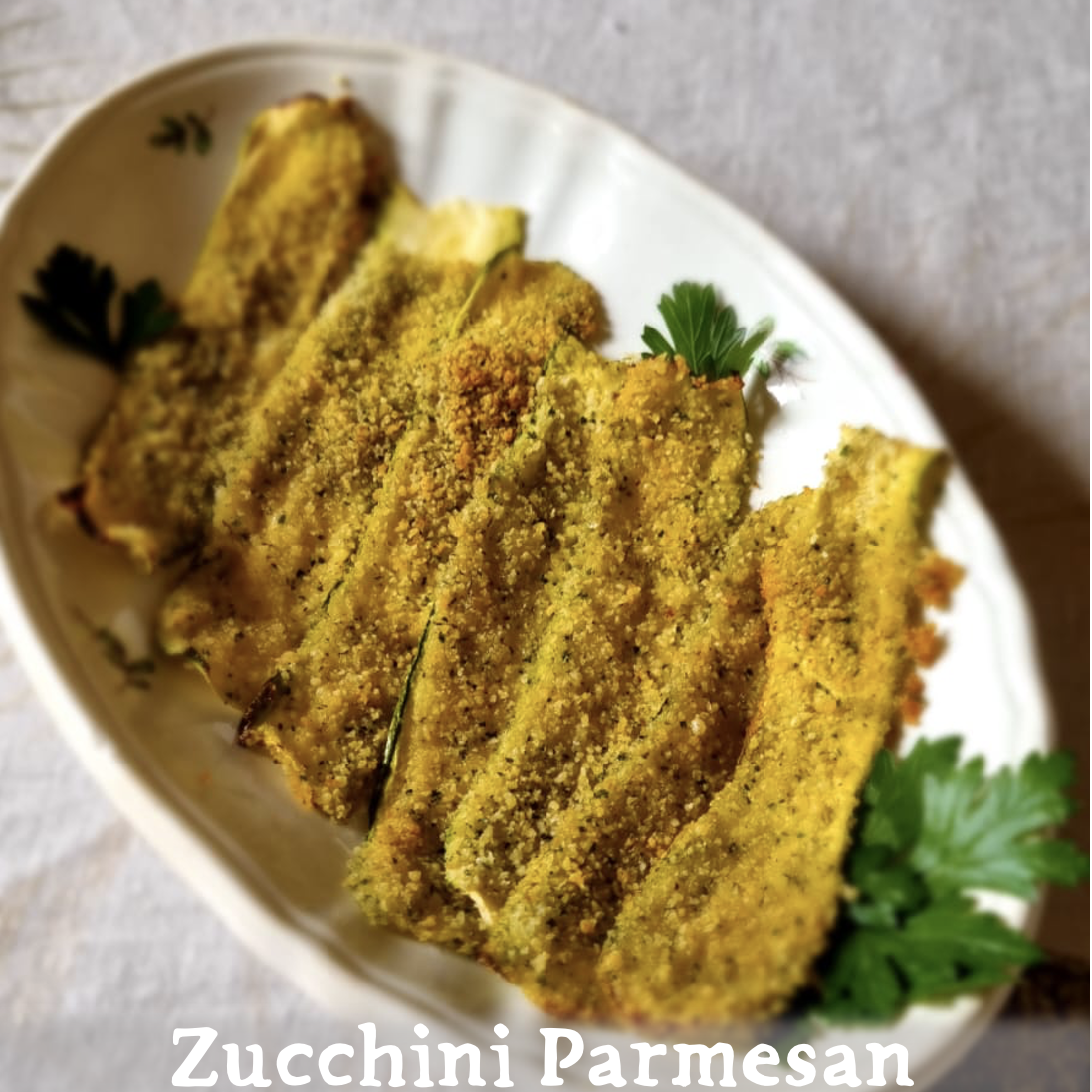 A white platter on a natural colored linen tablecloth, with seven thin slices of baked zucchini covered in breadcrumbs, garnished with parsley. With a folk lying across the zucchini. "Zucchini Parmesan" is written at the bottom.