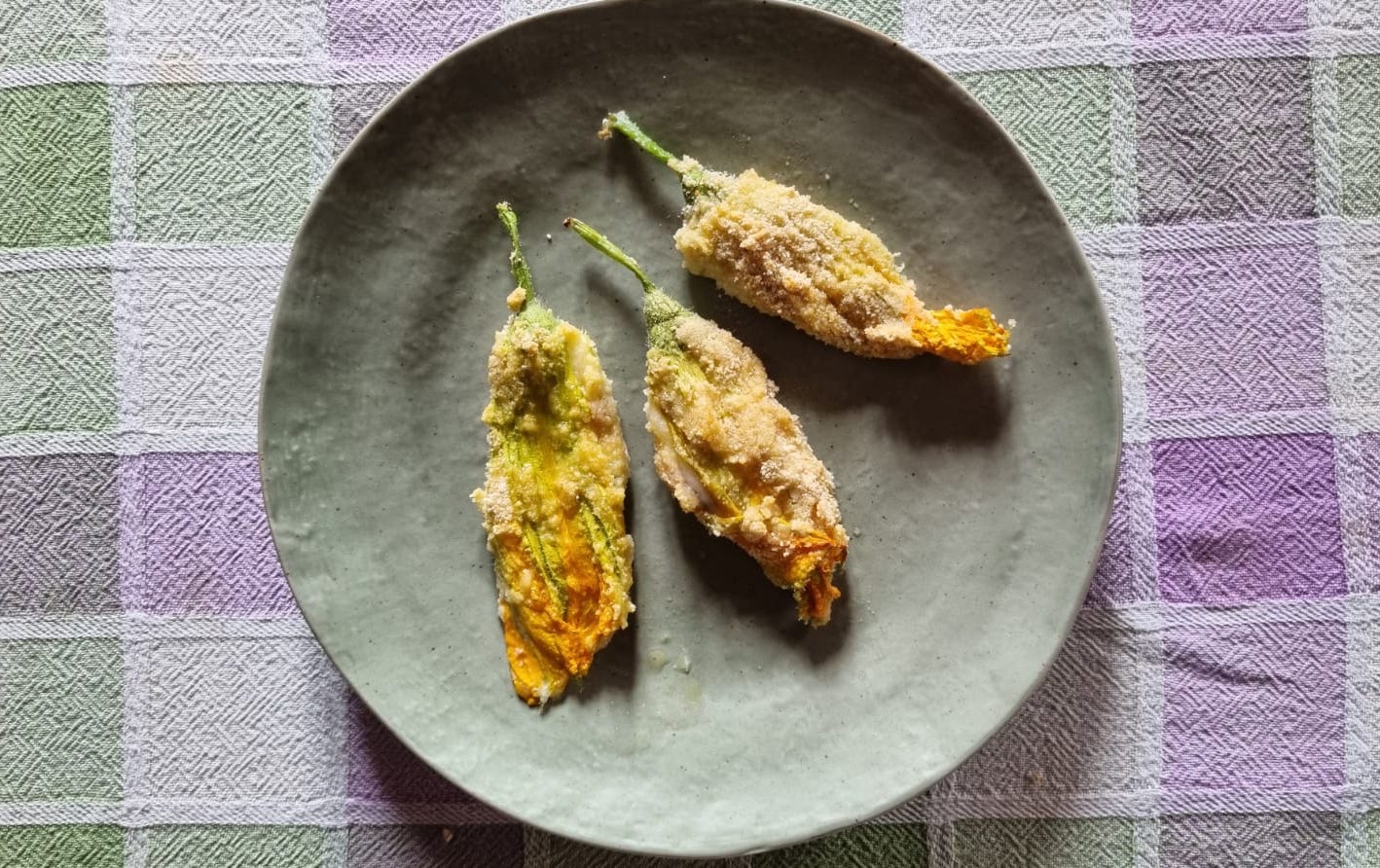 Three lightly breaded and baked vibrant zucchini flowers on a rustic gray ceramic plate, on a light green and lavender checked tablecloth. 