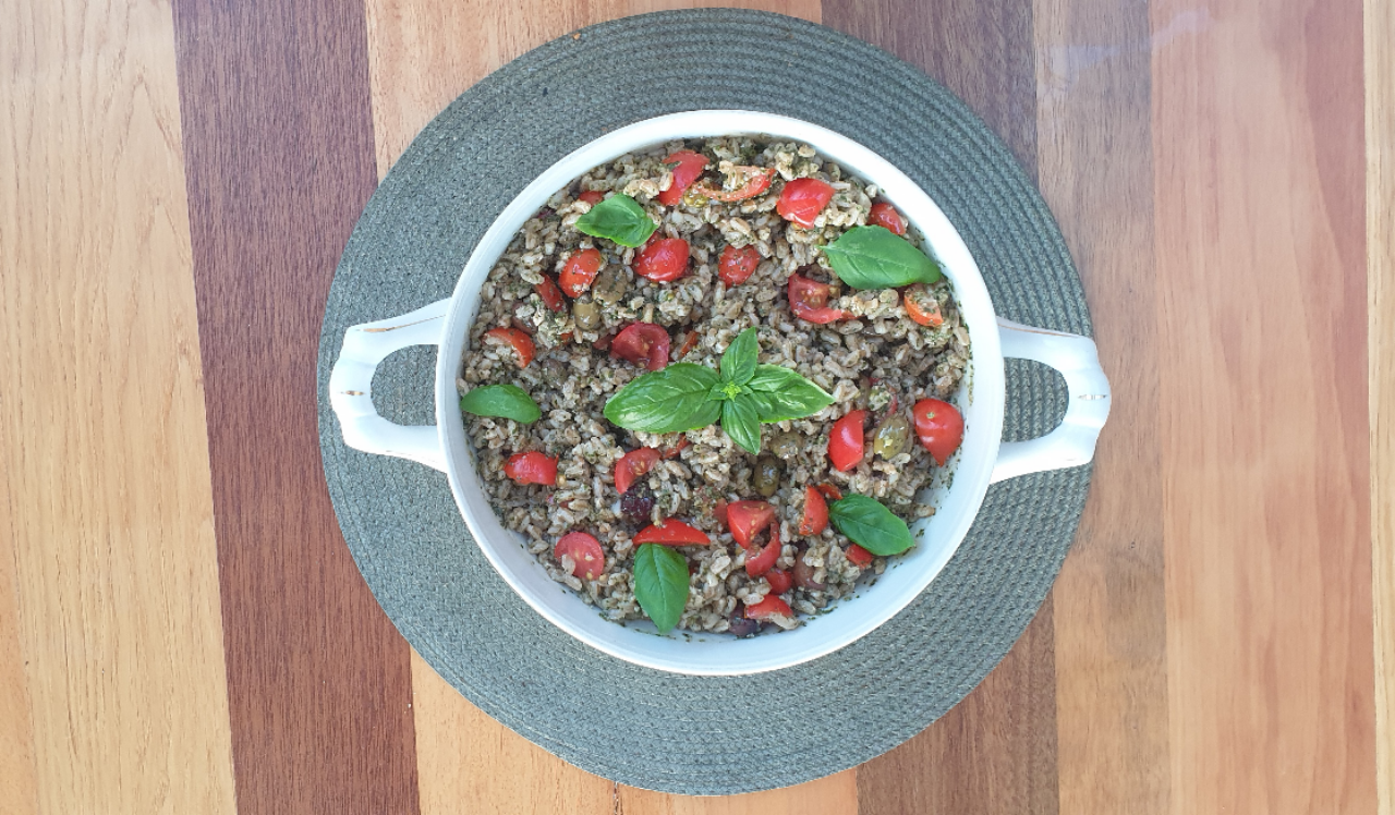 A white ceramic serving bowl filled with farro, chopped tomatoes, basil, and green olives. Garnished with fresh basil and set on a woven placemat and wood table.