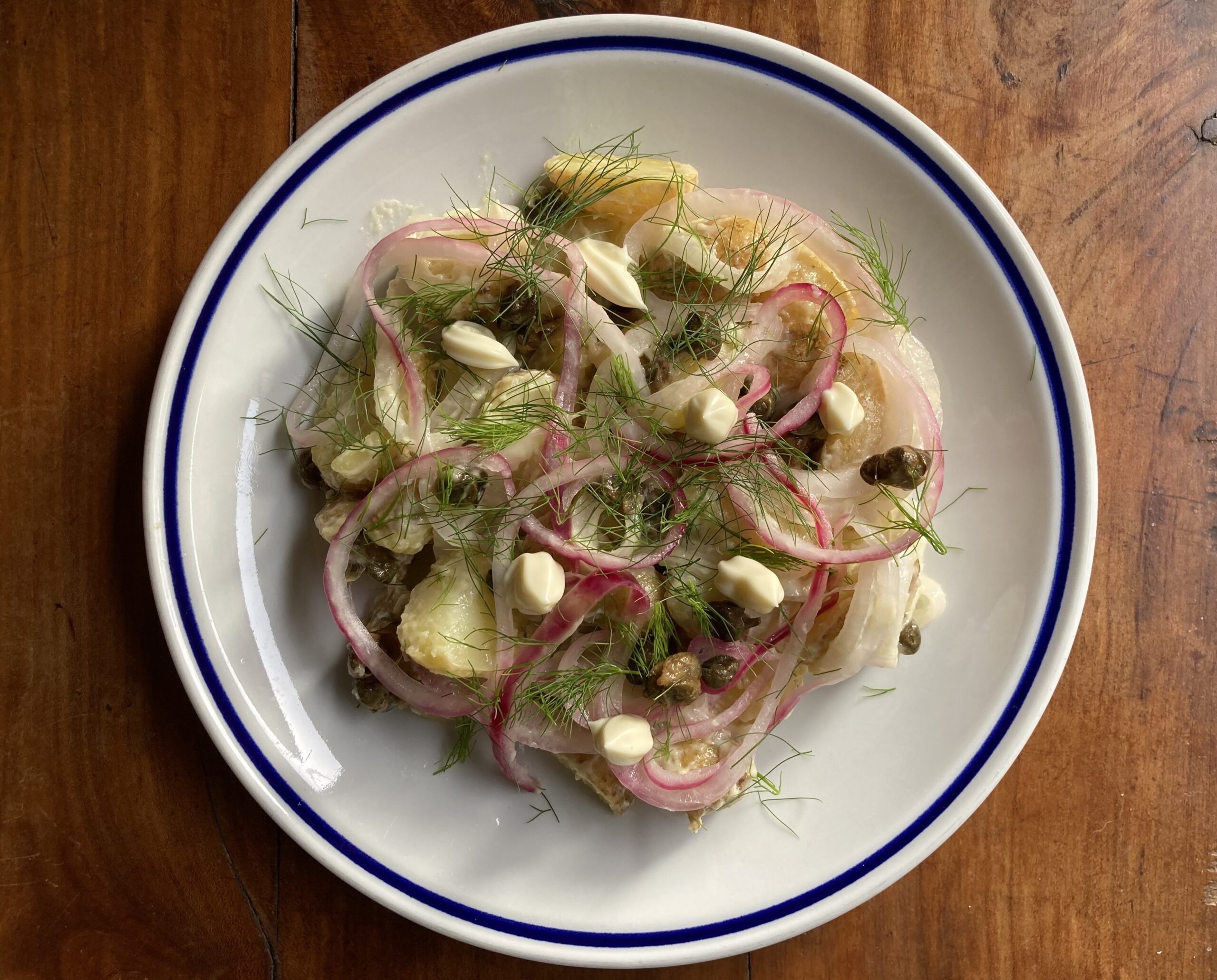 A white ceramic plate on wooden table surface, layer with wedges of potatoes, pickled red onions, capers, dollops of mayonnaise, and fresh fennel fronds.