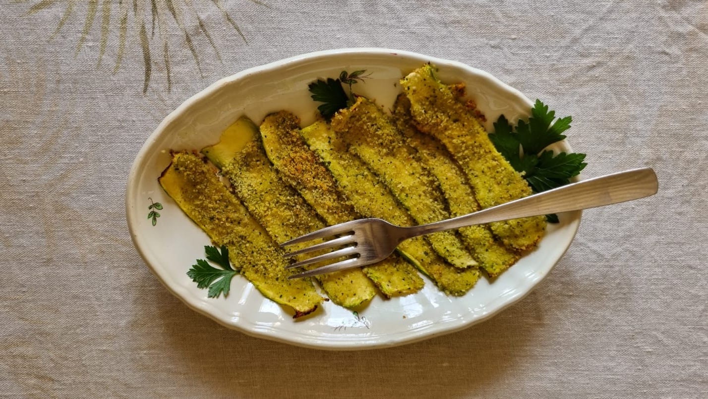 A white platter on a natural colored linen tablecloth, with seven thin slices of baked zucchini covered in breadcrumbs, garnished with parsley. With a folk lying across the zucchini.