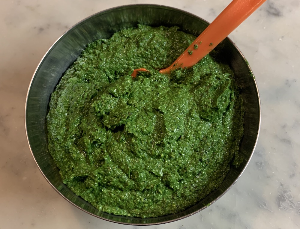 A metal bowl of deep green chunky pesto with a big orange spoon, on a marble tabletop.