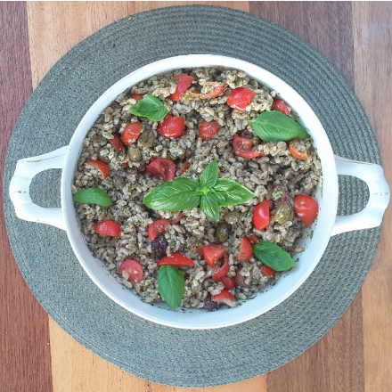 A white ceramic serving bowl filled with farro, chopped tomatoes, basil, and green olives. Garnished with fresh basil and set on a woven placemat.