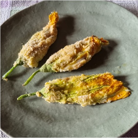 Three lightly breaded and baked vibrant zucchini flowers on a rustic gray ceramic plate.