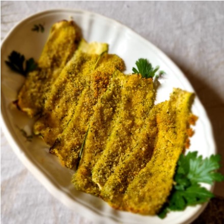 A white platter on a natural colored linen tablecloth, with seven thin slices of baked zucchini covered in breadcrumbs, garnished with parsley. 