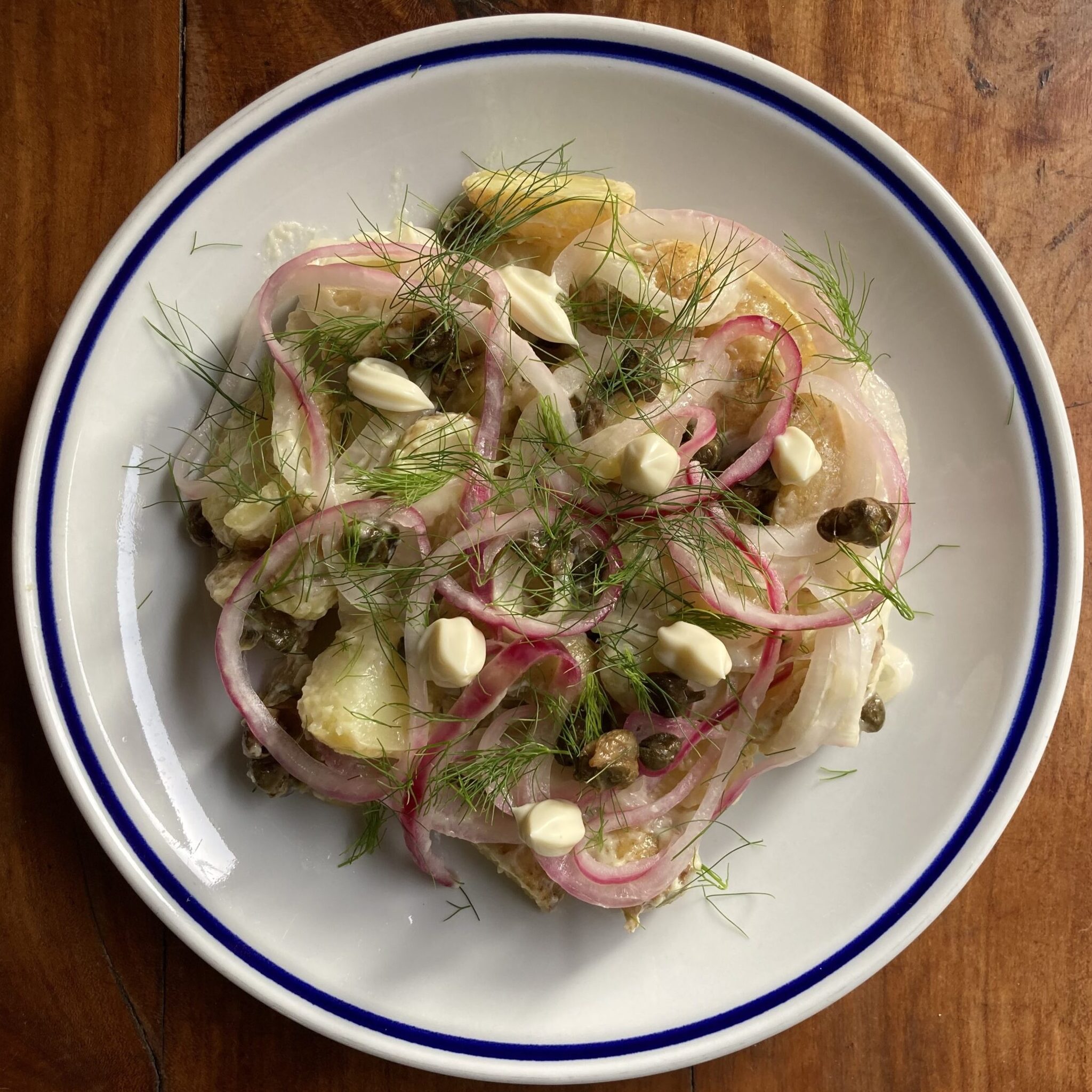 A white ceramic plate layered with wedges of potatoes, pickled red onions, capers, dollops of mayonnaise, and fresh fennel fronds.