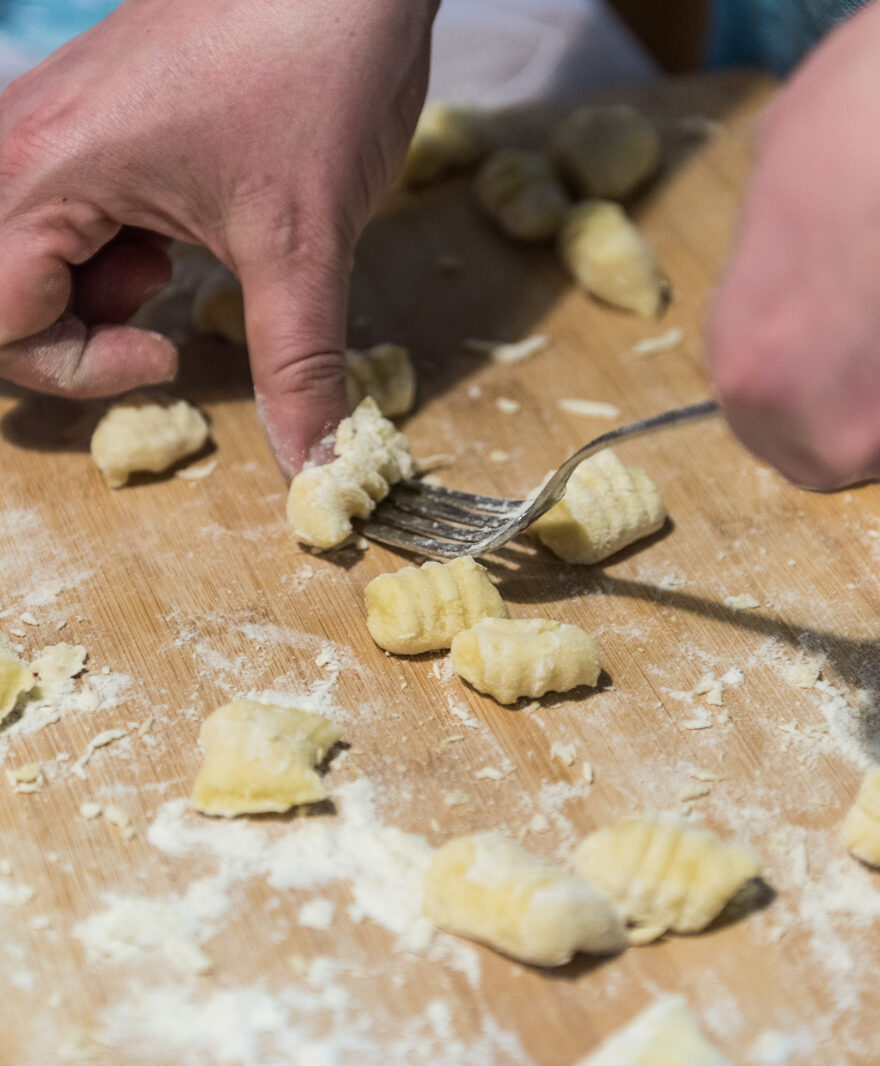 Close-up photo of two hands rolling gnocchi using a fork. Finished raw gnocchi surround, on a floured wooden cutting board.