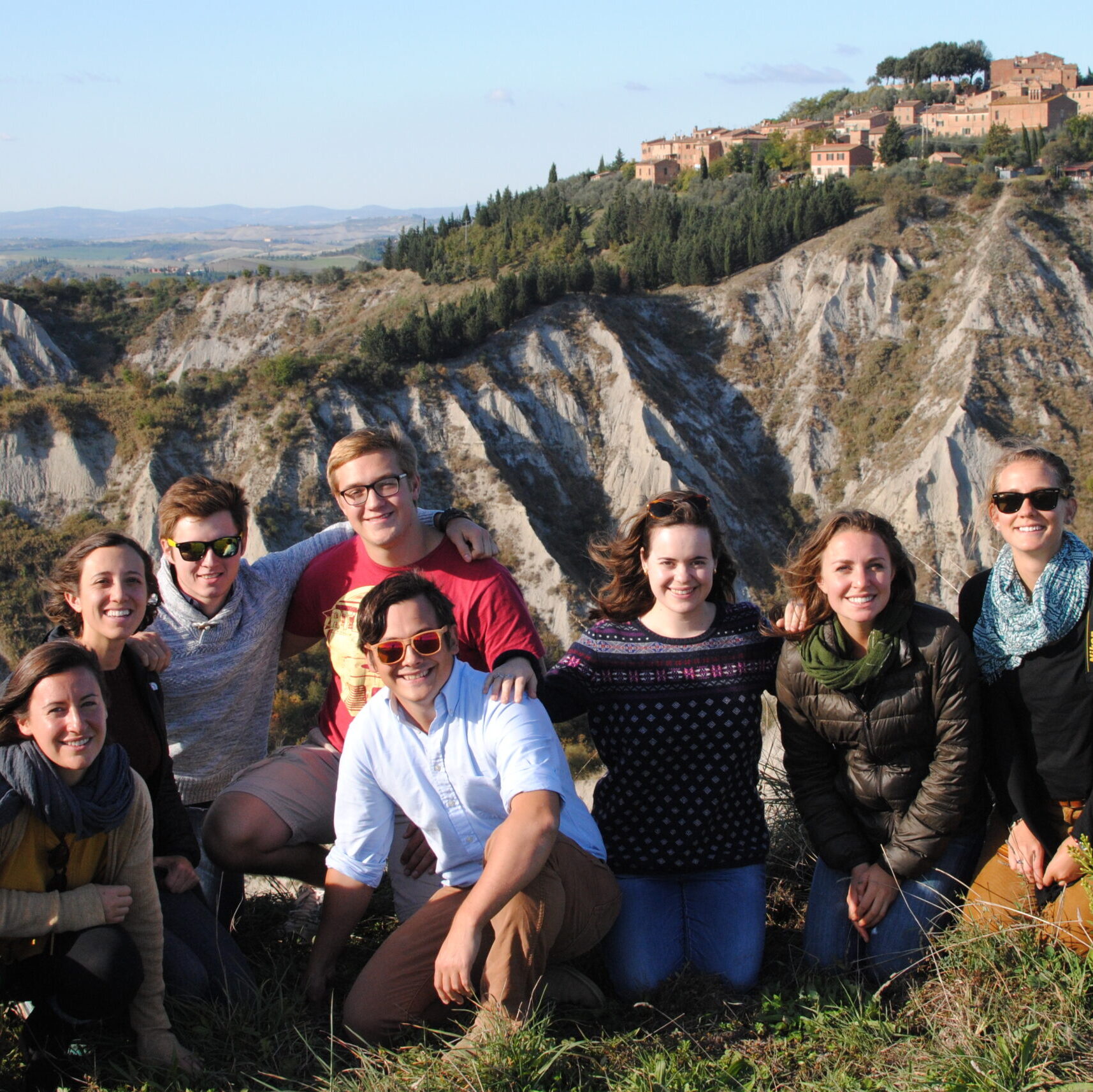 A group of interns kneeling together in a group, smiling at the camera. They are on a field trip and an old city on a mountain is in the background.