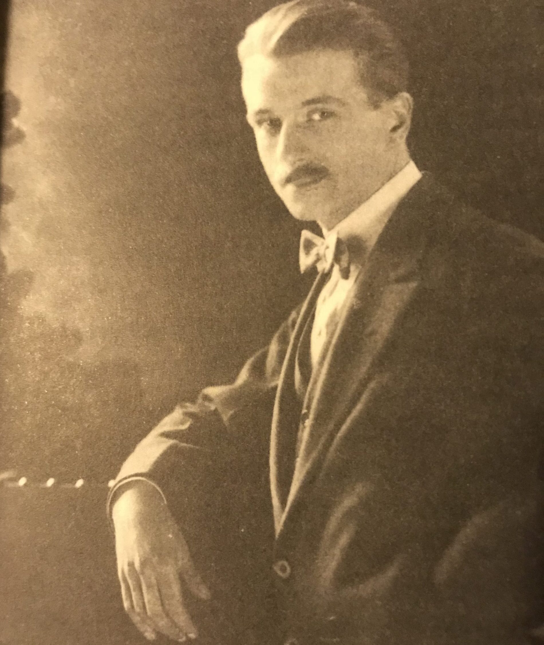 An old sepia toned photo of a man in a suit and bow tie looking at the camera. He has slicked back dark hair and a dark mustache.