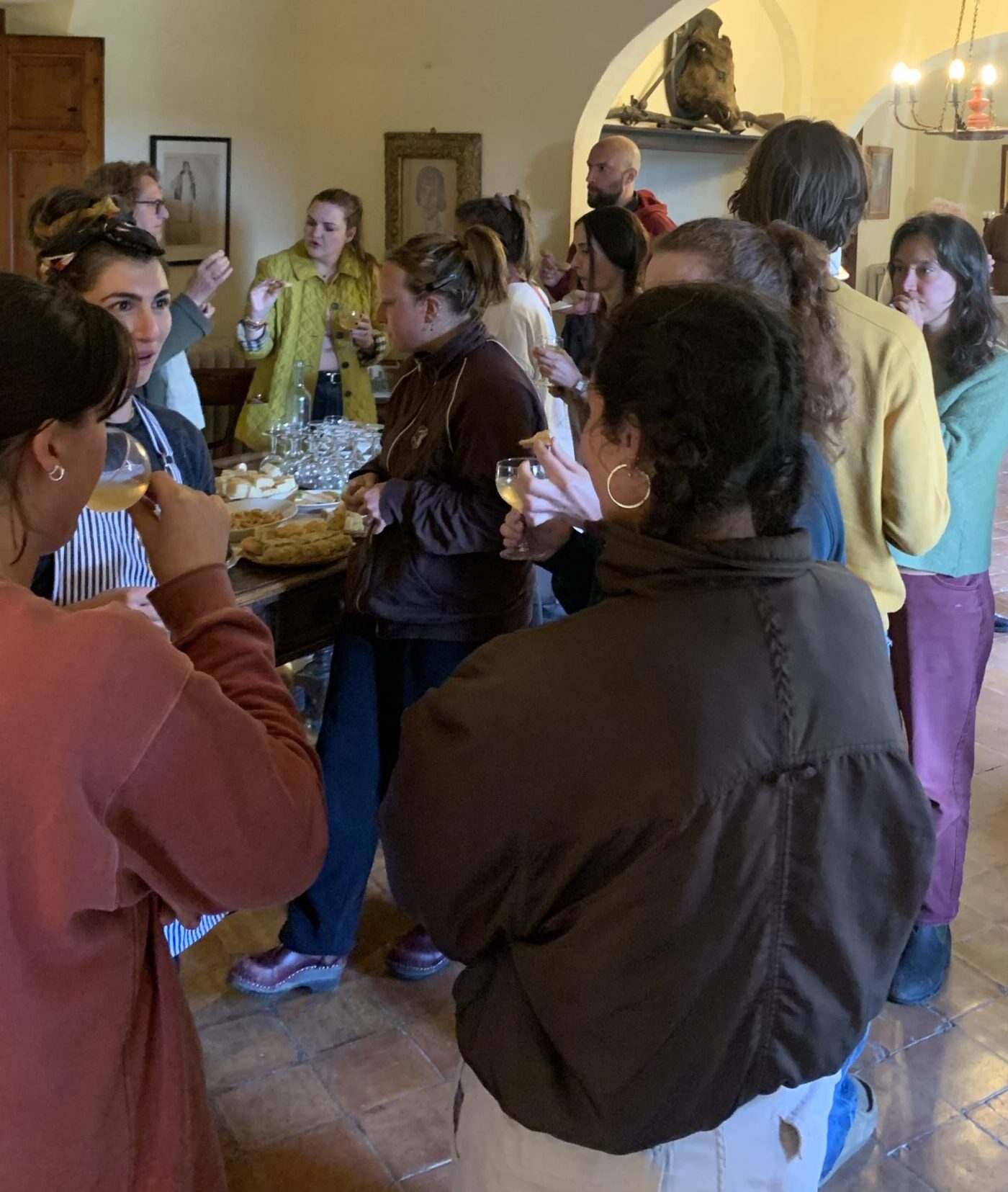 A candid photo of a group of people gathered in the Spannocchia dining room in small conversation groups. In the background, a table with food on it can be seen.