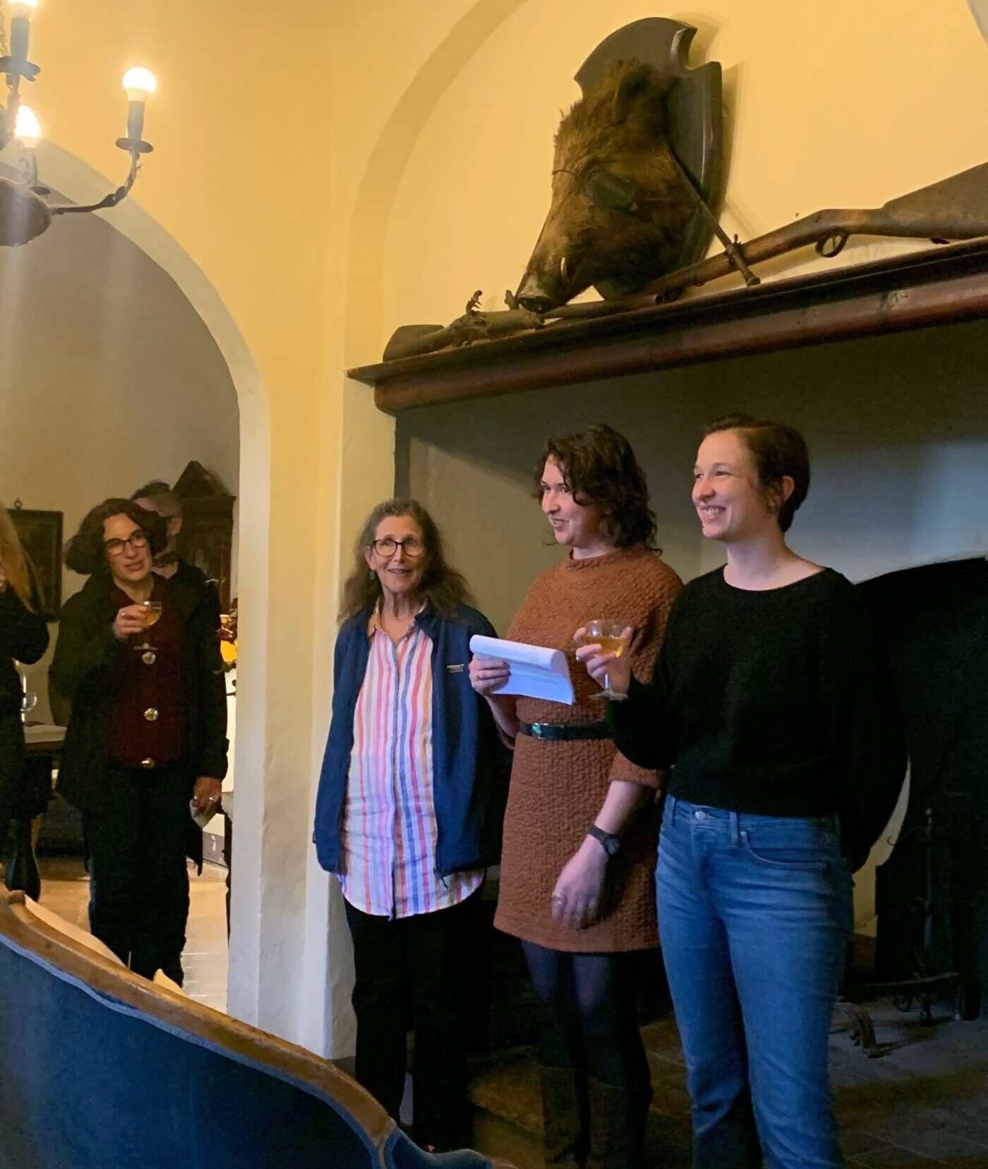 Four people standing in the Spannocchia living room. Three women stand together in front of a big fireplace, smiling out at the room. A wild boar's head is mounted on the wall above them.