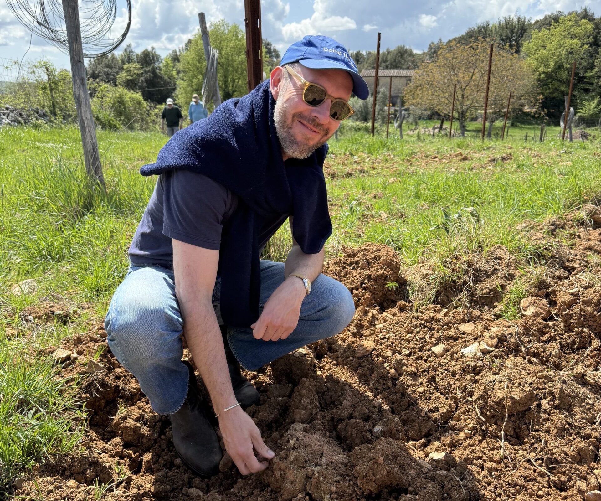 A man, Niccolò, squatting in the vineyard soil and smiling at the camera.