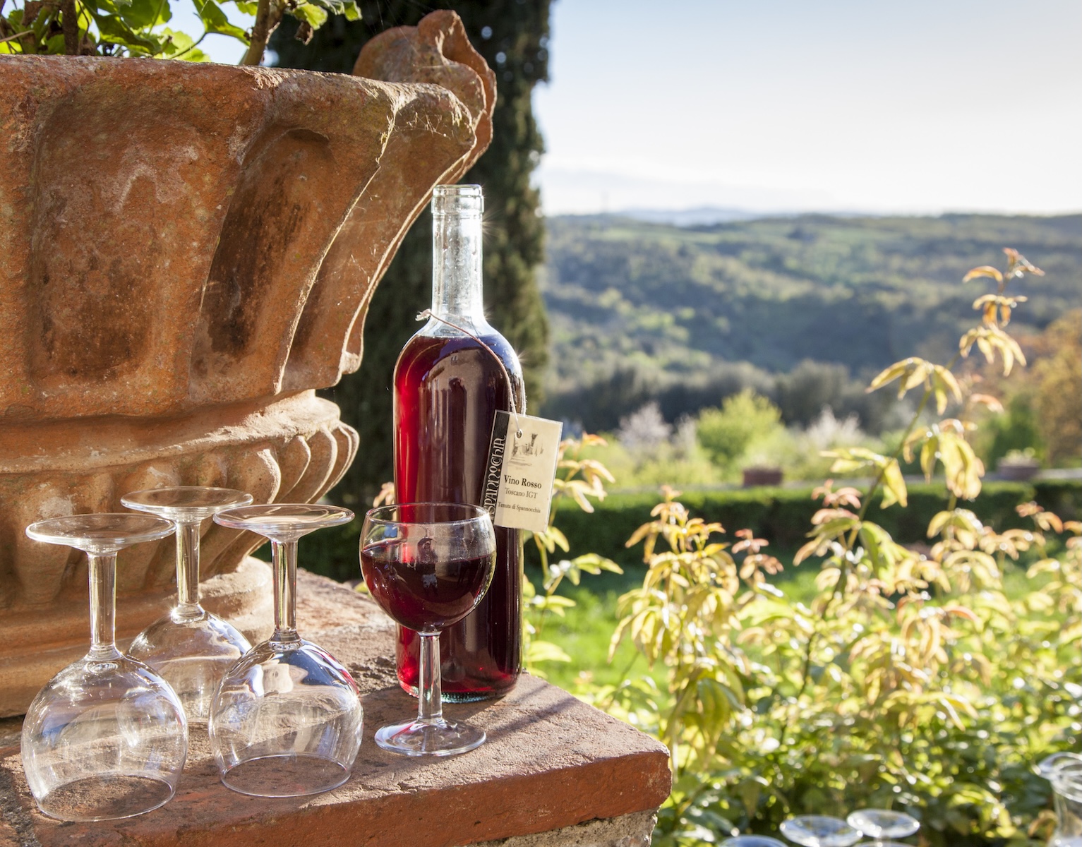 A bottle of wine and wine glasses on top of a terrace ledge, overlooking the villa lawn and Tuscan hills beyond.