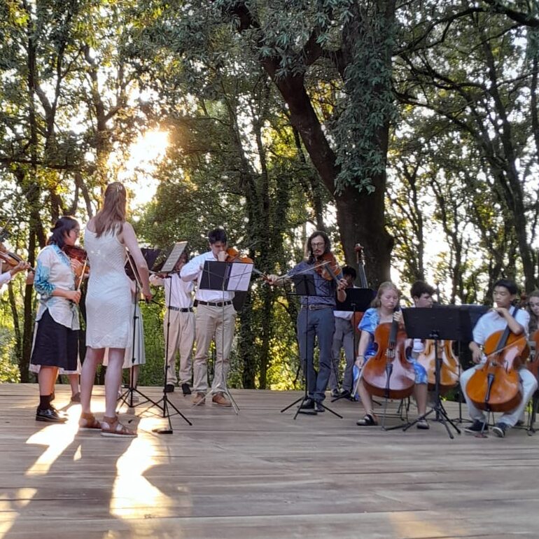 A photo of people standing on a wooden platform outside, surrounded by trees. They are playing string instruments as part of a chamber music concert.