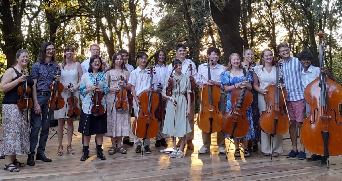 A photo of people standing on a wooden platform outside, surrounded by trees. They are smiling at the camera and holding their string instruments.