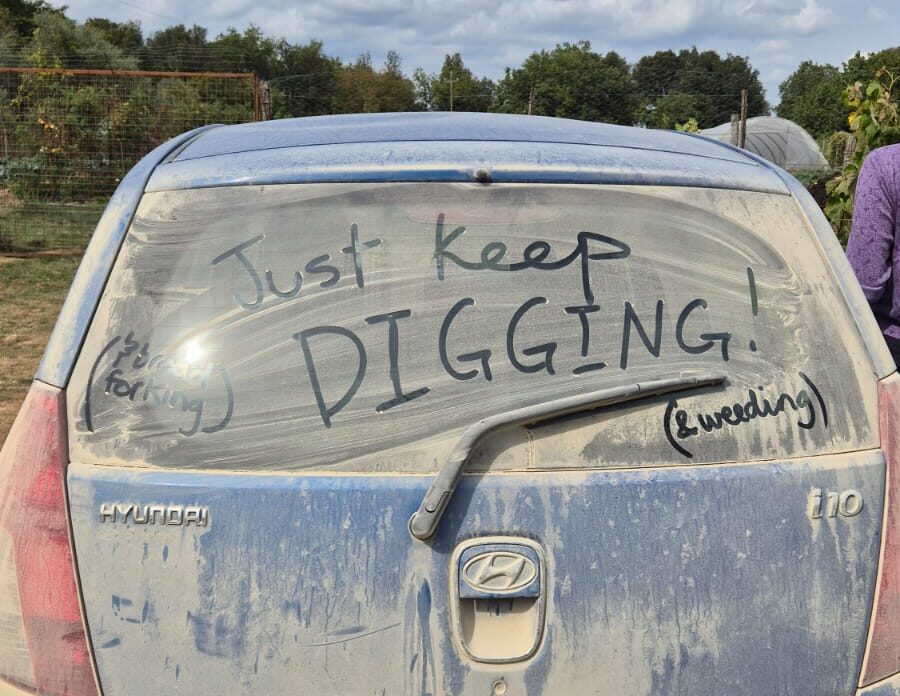 The back of a blue car used on the farm, covered in dust, with the words "Just keep digging!" "And broad forking" and "And weeding" written in the dust on the back window.